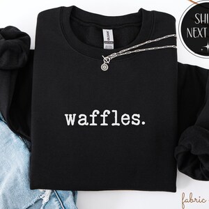 K&ouml;nnte beinhalten: Schwarzes Sweatshirt mit Rundhalsausschnitt und dem wei&szlig;en Aufdruck "waffles". Eine silberne Halskette mit einem kreisf&ouml;rmigen Anh&auml;nger liegt &uuml;ber dem Sweatshirt. Das Sweatshirt ist mit einer Jeans gefaltet.