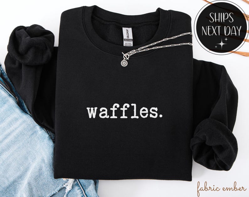 K&ouml;nnte beinhalten: Schwarzes Sweatshirt mit Rundhalsausschnitt und dem wei&szlig;en Aufdruck "waffles". Eine silberne Halskette mit einem kreisf&ouml;rmigen Anh&auml;nger liegt &uuml;ber dem Sweatshirt. Das Sweatshirt ist mit einer Jeans gefaltet.