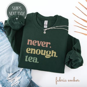 以下が含まれることがあります： ピンク、ベージュ、緑色の「never. enough. tea.」というフレーズが入った濃い緑色のスウェットシャツ。ペンダント付きのシルバーネックレスがスウェットシャツの上にあります。「ships next day」という言葉が左上の円の中にあります。