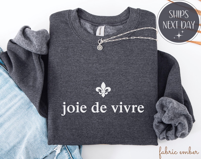 May include: A charcoal grey sweatshirt featuring the words "joie de vivre" in white, and a white fleur-de-lis. A silver necklace is draped across the garment. The sleeves are turned up, showing a lighter grey lining.
