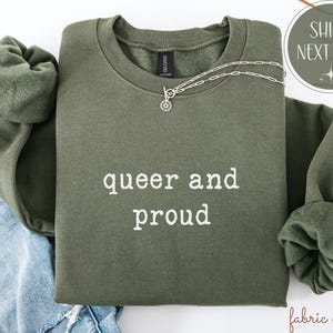 May include: An olive green sweatshirt with the words "queer and proud" in white lettering. A silver necklace with a circular pendant rests on the sweatshirt. The sweatshirt is folded on top of blue denim.