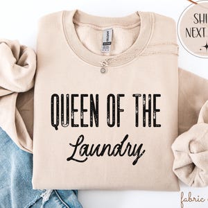 May include: A beige sweatshirt with the text "QUEEN OF THE LAUNDRY" in black. The sweatshirt is folded with a necklace and a pair of jeans visible. The image also includes a "SHIPS NEXT DAY" graphic.