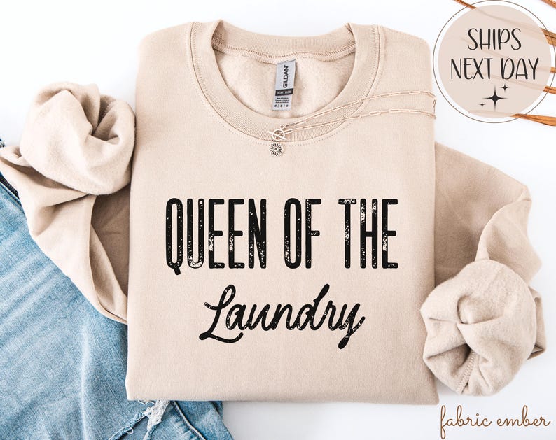 May include: A beige sweatshirt with the text "QUEEN OF THE LAUNDRY" in black. The sweatshirt is folded with a necklace and a pair of jeans visible. The image also includes a "SHIPS NEXT DAY" graphic.