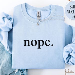 May include: A light blue sweatshirt with the word "nope" printed in black. A silver necklace with a small pendant is on the sweatshirt. The sleeves are folded. The text "fabric ember" is in the bottom right corner.