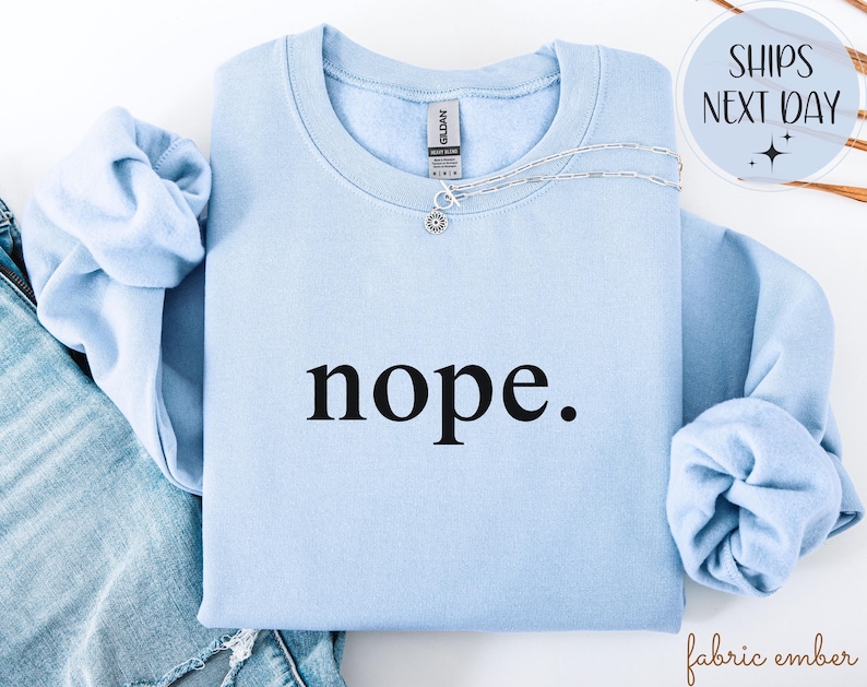 May include: A light blue sweatshirt with the word "nope" printed in black. A silver necklace with a small pendant is on the sweatshirt. The sleeves are folded. The text "fabric ember" is in the bottom right corner.