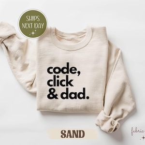 May include: Sand-coloured sweatshirt with the text "code, click & dad" in black. The sweatshirt has a crew neck and long sleeves with rolled cuffs. The word "SAND" is printed on a tan banner.