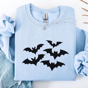 Puede incluir: Una sudadera azul claro con un dise&ntilde;o de murci&eacute;lagos negros. La sudadera de cuello redondo est&aacute; doblada y se muestra con un collar plateado y un par de jeans azules. Los murci&eacute;lagos est&aacute;n en varias posiciones.