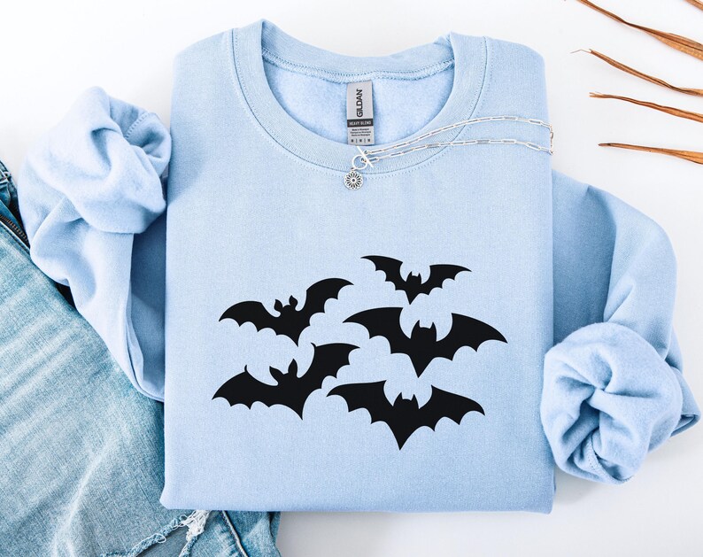 Puede incluir: Una sudadera azul claro con un dise&ntilde;o de murci&eacute;lagos negros. La sudadera de cuello redondo est&aacute; doblada y se muestra con un collar plateado y un par de jeans azules. Los murci&eacute;lagos est&aacute;n en varias posiciones.