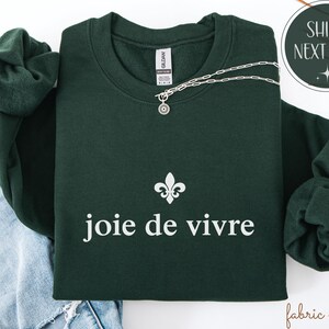 May include: A dark green sweatshirt featuring the words "joie de vivre" in white, and a fleur-de-lis symbol. A silver necklace with a small pendant is draped across the sweatshirt. The sweatshirt is folded on top of a pair of blue jeans.