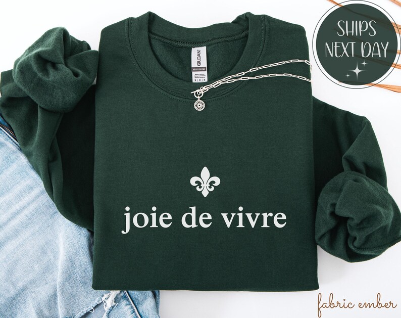 May include: A dark green sweatshirt featuring the words "joie de vivre" in white, and a fleur-de-lis symbol. A silver necklace with a small pendant is draped across the sweatshirt. The sweatshirt is folded on top of a pair of blue jeans.