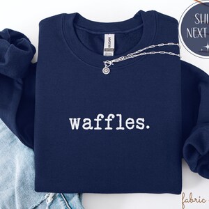 K&ouml;nnte beinhalten: Ein marineblaues Sweatshirt mit dem Wort "waffles." in Wei&szlig; aufgedruckt. Eine silberne Halskette mit einem kreisf&ouml;rmigen Anh&auml;nger liegt &uuml;ber dem Sweatshirt. Das Sweatshirt ist auf einer Jeans gefaltet.