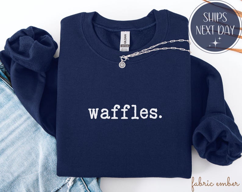K&ouml;nnte beinhalten: Ein marineblaues Sweatshirt mit dem Wort "waffles." in Wei&szlig; aufgedruckt. Eine silberne Halskette mit einem kreisf&ouml;rmigen Anh&auml;nger liegt &uuml;ber dem Sweatshirt. Das Sweatshirt ist auf einer Jeans gefaltet.