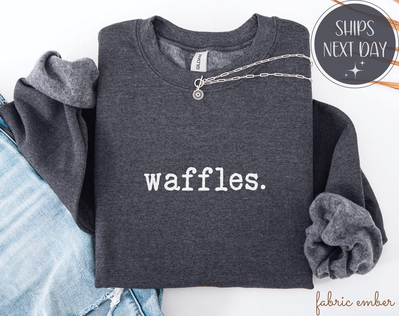 K&ouml;nnte beinhalten: Ein dunkelgraues Sweatshirt mit dem Wort "waffles" in Wei&szlig; aufgedruckt. Eine silberne Halskette mit Anh&auml;nger liegt &uuml;ber dem Shirt. Die &Auml;rmel sind umgeschlagen und zeigen ein helleres graues Futter. Das Shirt liegt auf einer Jeans.