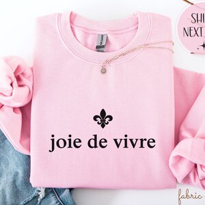 May include: A pale pink sweatshirt featuring the words "joie de vivre" in black, with a black fleur-de-lis symbol. A gold necklace with a small pendant is placed on the folded garment. The sweatshirt is folded on top of blue jeans.