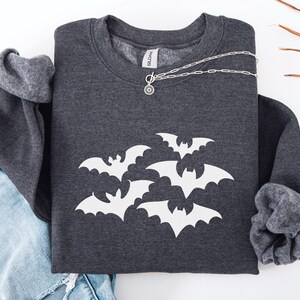 Puede incluir: Sudadera gris oscuro con un dise&ntilde;o de murci&eacute;lagos blancos. Un collar plateado con un colgante circular descansa sobre la sudadera. Las mangas est&aacute;n dobladas y se ve un par de vaqueros azules. La sudadera es de la marca Gildan.