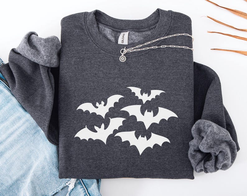 Puede incluir: Sudadera gris oscuro con un dise&ntilde;o de murci&eacute;lagos blancos. Un collar plateado con un colgante circular descansa sobre la sudadera. Las mangas est&aacute;n dobladas y se ve un par de vaqueros azules. La sudadera es de la marca Gildan.