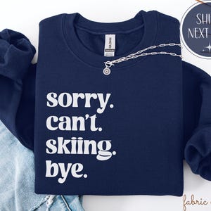 May include: Navy blue sweatshirt with the text "sorry. can't. skiing. bye." in white. A silver necklace with a circular pendant is draped across the front. The sweatshirt is folded on top of a pair of blue jeans.