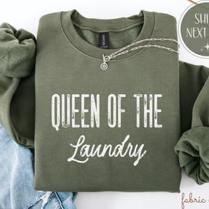 May include: An olive green sweatshirt with the text "QUEEN OF THE LAUNDRY" in white. A silver necklace with a pendant is draped across the front. The sweatshirt is folded on top of a pair of blue jeans.