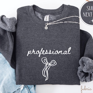 Può includere: Una felpa grigio scuro con la scritta "professional" in corsivo bianco e un fiocco bianco. Una collana d'argento con un ciondolo è drappeggiata sulla felpa. La felpa è piegata sopra un paio di jeans blu.