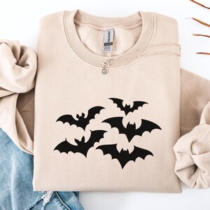 Puede incluir: Sudadera beige con un dise&ntilde;o de murci&eacute;lagos negros. Los murci&eacute;lagos est&aacute;n agrupados, con las alas extendidas. La sudadera tiene cuello redondo y mangas largas. Se ve un collar plateado con un peque&ntilde;o colgante.