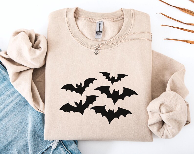 Puede incluir: Sudadera beige con un dise&ntilde;o de murci&eacute;lagos negros. Los murci&eacute;lagos est&aacute;n agrupados, con las alas extendidas. La sudadera tiene cuello redondo y mangas largas. Se ve un collar plateado con un peque&ntilde;o colgante.