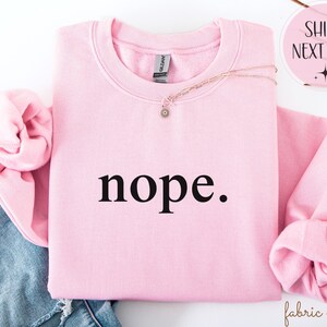 May include: A light pink sweatshirt with the word "nope." printed in black. The sweatshirt has a crew neck and long sleeves. A gold necklace with a small pendant is draped across the front.