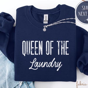 May include: A navy blue sweatshirt with the text "QUEEN OF THE Laundry" in white. A silver necklace with a pendant is draped across the front. The sweatshirt is folded on top of a pair of blue jeans.