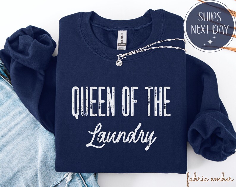 May include: A navy blue sweatshirt with the text "QUEEN OF THE Laundry" in white. A silver necklace with a pendant is draped across the front. The sweatshirt is folded on top of a pair of blue jeans.