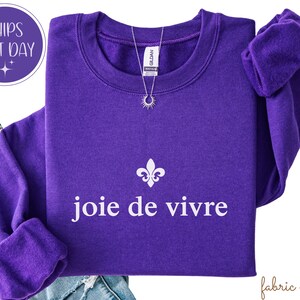 May include: A vibrant purple sweatshirt featuring the words "joie de vivre" in white, and a fleur-de-lis symbol. A silver necklace with a sun pendant is on the sweatshirt. The sweatshirt is folded over a pair of blue jeans.