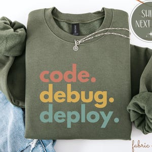 May include: An olive green sweatshirt with the words "code. debug. deploy." in coral, yellow, and teal. A silver necklace with a small pendant is draped across the front. The sweatshirt is folded on top of blue denim.