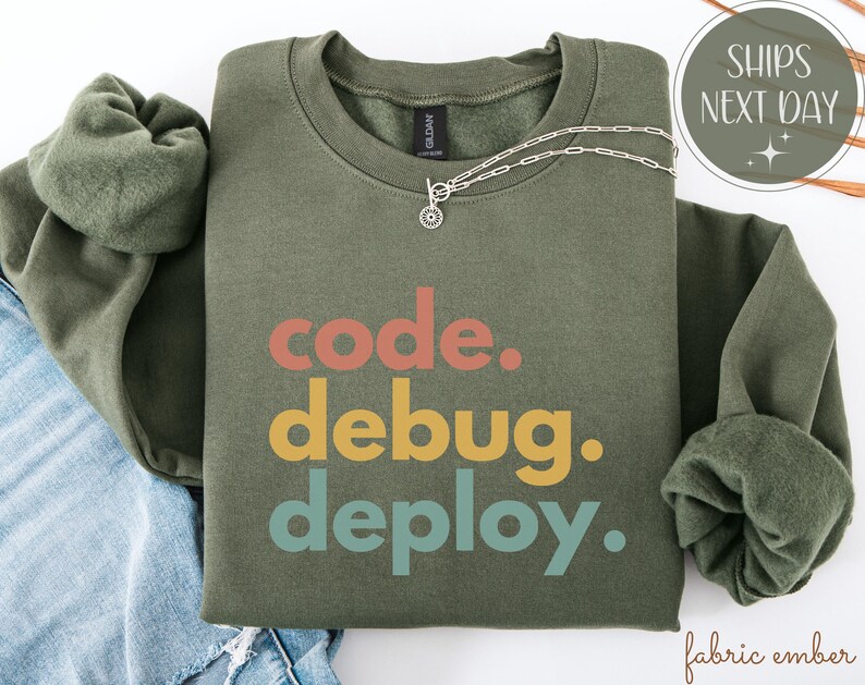 May include: An olive green sweatshirt with the words "code. debug. deploy." in coral, yellow, and teal. A silver necklace with a small pendant is draped across the front. The sweatshirt is folded on top of blue denim.