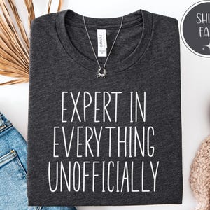 May include: Dark gray t-shirt with the text "EXPERT IN EVERYTHING UNOFFICIALLY" in white. A silver necklace with a sunburst pendant is on the shirt. The shirt is folded on a white surface with a pair of blue jeans.
