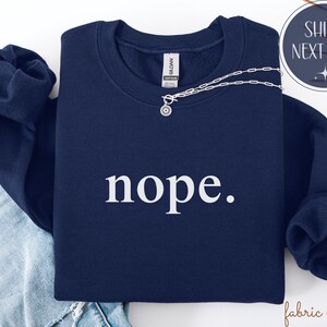 May include: A navy blue sweatshirt with the word "nope" in white, displayed on a folded garment. A silver necklace with a circular pendant rests on the sweatshirt. The image also includes a pair of blue jeans.