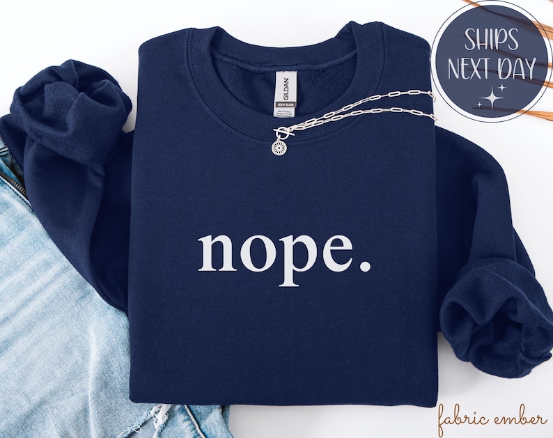 May include: A navy blue sweatshirt with the word "nope" in white, displayed on a folded garment. A silver necklace with a circular pendant rests on the sweatshirt. The image also includes a pair of blue jeans.