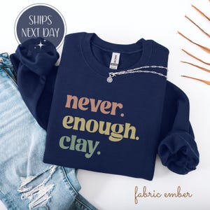 May include: Navy blue sweatshirt with the text "never. enough. clay." in peach, yellow-green, and green. A silver necklace with a pendant is on the sweatshirt. The words "ships next day" are in a circle in the upper left corner.
