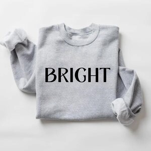May include: A pale grey sweatshirt featuring the word "BRIGHT" in large, black capital letters. The sleeves are rolled up, and the crew neck is visible. The garment is made of a soft material, suitable for casual wear.