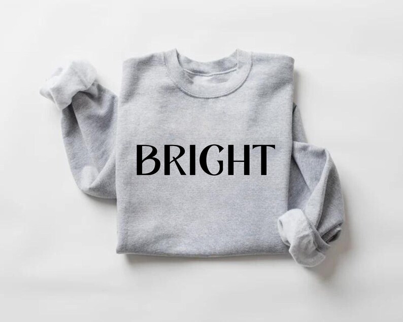 May include: A pale grey sweatshirt featuring the word "BRIGHT" in large, black capital letters. The sleeves are rolled up, and the crew neck is visible. The garment is made of a soft material, suitable for casual wear.