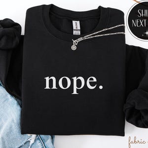 May include: Black sweatshirt with the word "nope." in white, a silver necklace, and a black hoodie. The sweatshirt is a crew neck style. The text is centered on the front of the shirt. The image also includes a denim jacket.