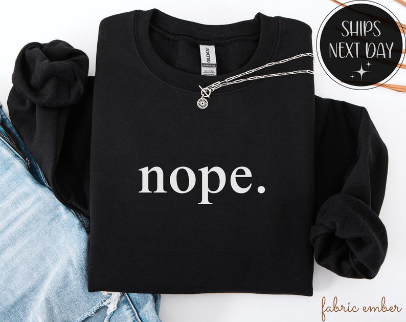 May include: Black sweatshirt with the word "nope." in white, a silver necklace, and a black hoodie. The sweatshirt is a crew neck style. The text is centered on the front of the shirt. The image also includes a denim jacket.