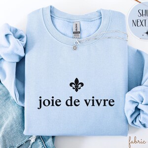 May include: A pale blue sweatshirt with the words "joie de vivre" in black. A silver necklace with a small pendant is on the sweatshirt. A black fleur-de-lis symbol is above the text.
