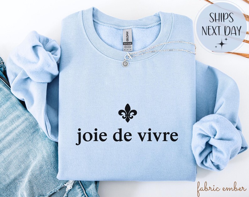 May include: A pale blue sweatshirt with the words "joie de vivre" in black. A silver necklace with a small pendant is on the sweatshirt. A black fleur-de-lis symbol is above the text.