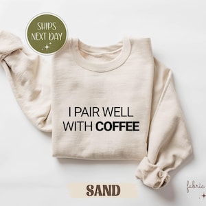 May include: A sand-coloured sweatshirt with the text "I Pair Well With Coffee" in black. The long-sleeved top is folded, with the sleeves cuffed. The word "SAND" is printed in a tan rectangle at the bottom. The brand name "fabric ember" is printed in the bottom right corner.