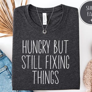 May include: Dark gray t-shirt with the text "HUNGRY BUT STILL FIXING THINGS" in white. A silver necklace with a sunburst pendant is on the shirt. The shirt is folded on a white surface next to a pair of blue jeans.