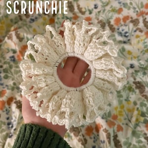 Può includere: Un elastico per capelli color crema con effetto pizzo. L'elastico è realizzato in materiale all'uncinetto e ha un bordo a balze. Il testo "LACE-EFFECT SCRUNCHIE" e "PDF PATTERN" sono visibili nell'immagine.