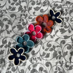 May include: Five crocheted flower hair scrunchies in different colours: navy blue, teal, pink, orange, and brown. Each flower has a white crocheted border.