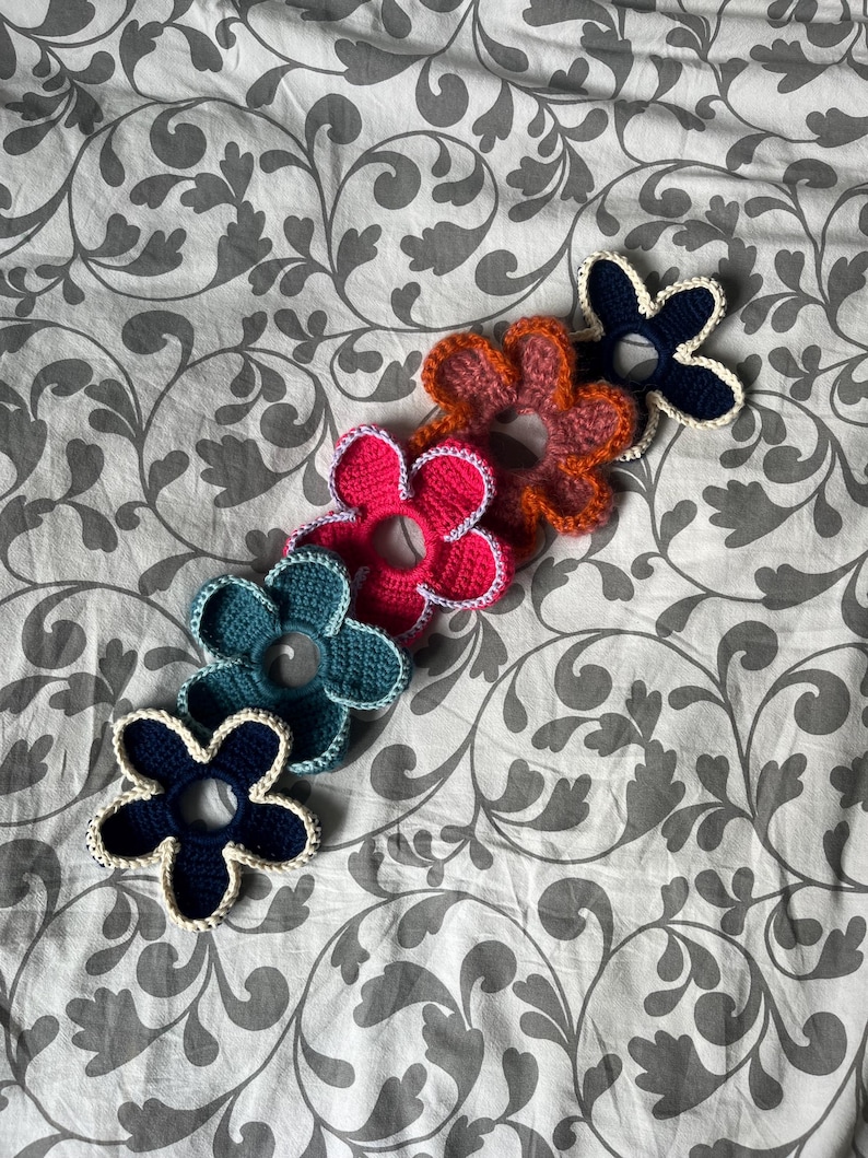 May include: Five crocheted flower hair scrunchies in different colours: navy blue, teal, pink, orange, and brown. Each flower has a white crocheted border.