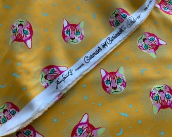May include: A bright yellow fabric featuring a repeating pattern of pink and orange cat faces with teal eyes, surrounded by light blue crescent moons and lightning bolts. The white selvage edge displays the text Free Spirit presents Curiouser and Curiouser by Tula Pink.