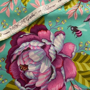 Op de afbeelding: Een turquoise stof met een grote paarse en roze pioenroos, groene bladeren en kleine bijen. De witte zelfkant toont de tekst: Moon Garden presents by Tula Pink PWTP 195 Kabloom. Het bloemmotief herhaalt zich over de gehele turquoise achtergrond.