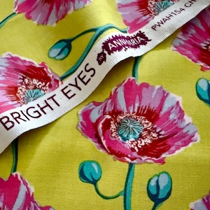 Könnte beinhalten: Nahaufnahme eines Stoffes mit einem sich wiederholenden Blumenmuster aus rosa Mohnblumen auf gelbem Grund. Die Blüten haben blaue Zentren. Ein weißes Band mit dem Text "BRIGHT EYES" und "ANNMARIA" ist sichtbar.