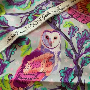 Op de afbeelding: Een lichtgrijze stof met een levendige uilenillustratie in roze, paarse en oranje tinten, omringd door gestileerd paars en groen gebladerte en kleine sterren. De zelfkant toont de tekst Moon Garden by Tula Pink Free Spirit.